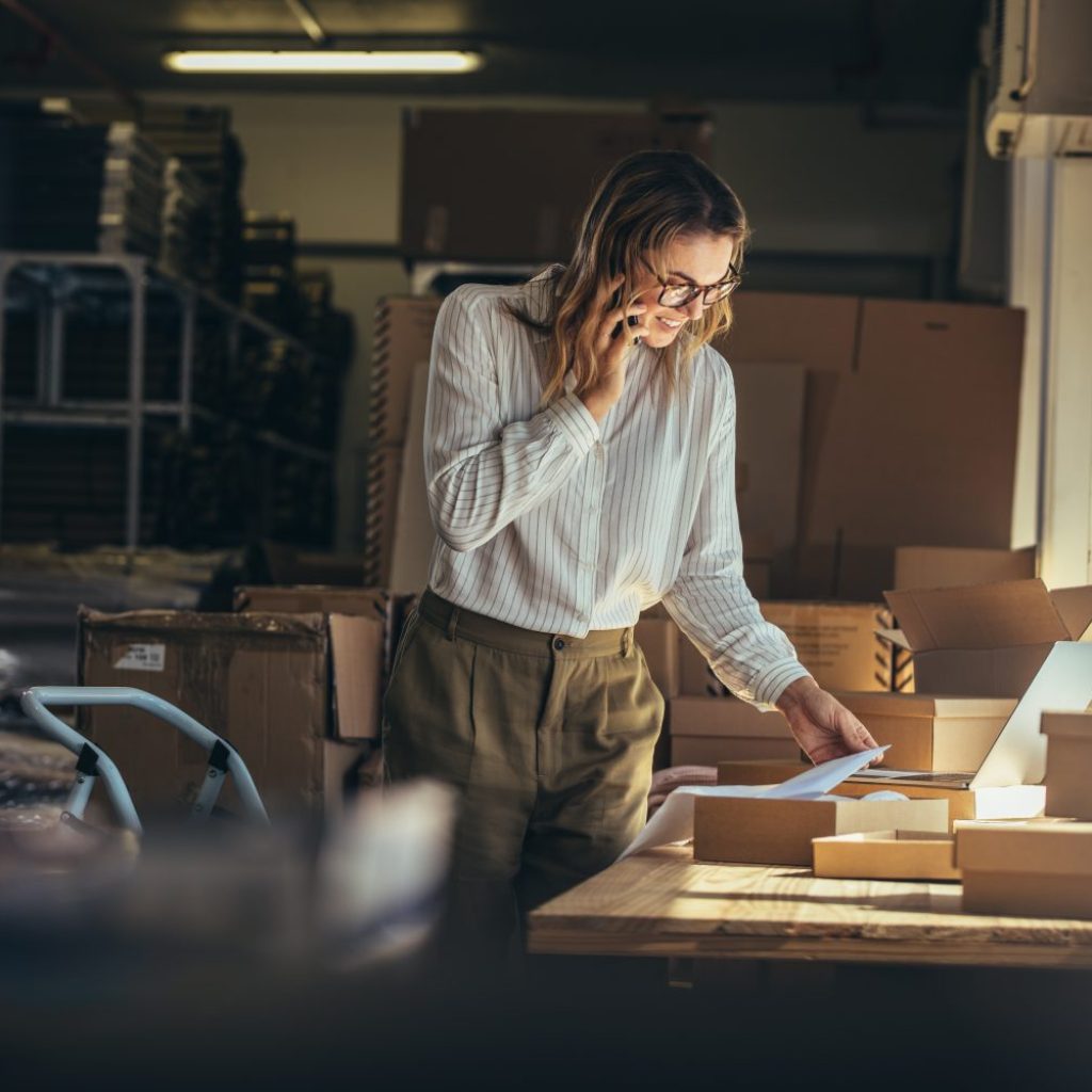 Woman online seller confirming orders from customer on the phone. E-commerce business owner looking at the papers and talking on phone in store warehouse.